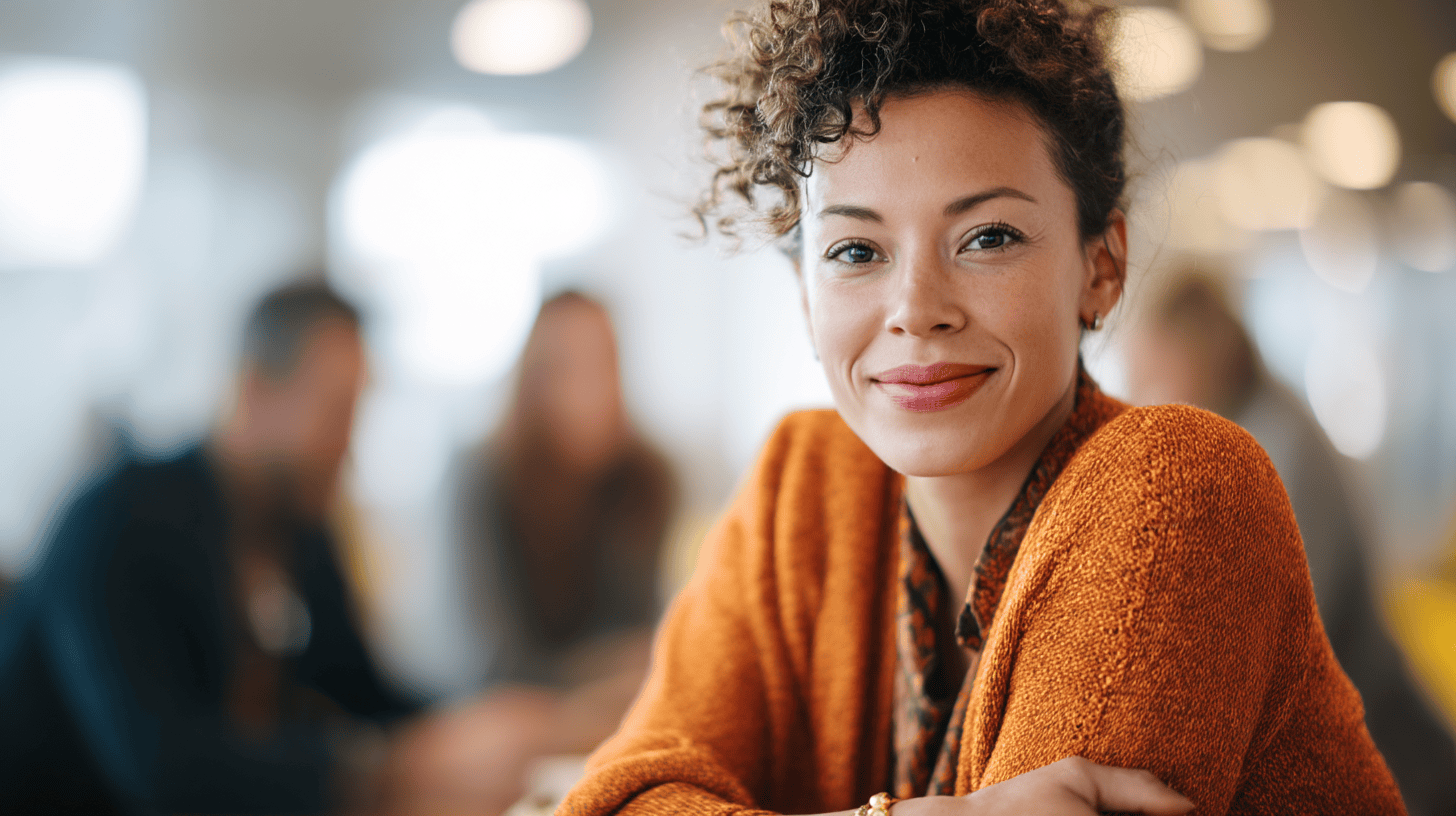 Smiling woman with curly hair wearing an orange sweater, seated at a table with blurred background figures.