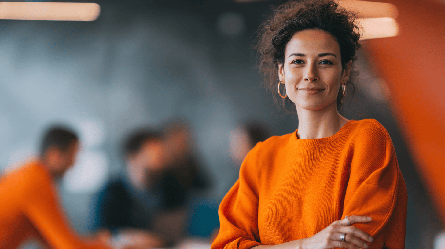 Woman in an orange sweater stands confidently, smiling, with a blurred background of people in a meeting setting.