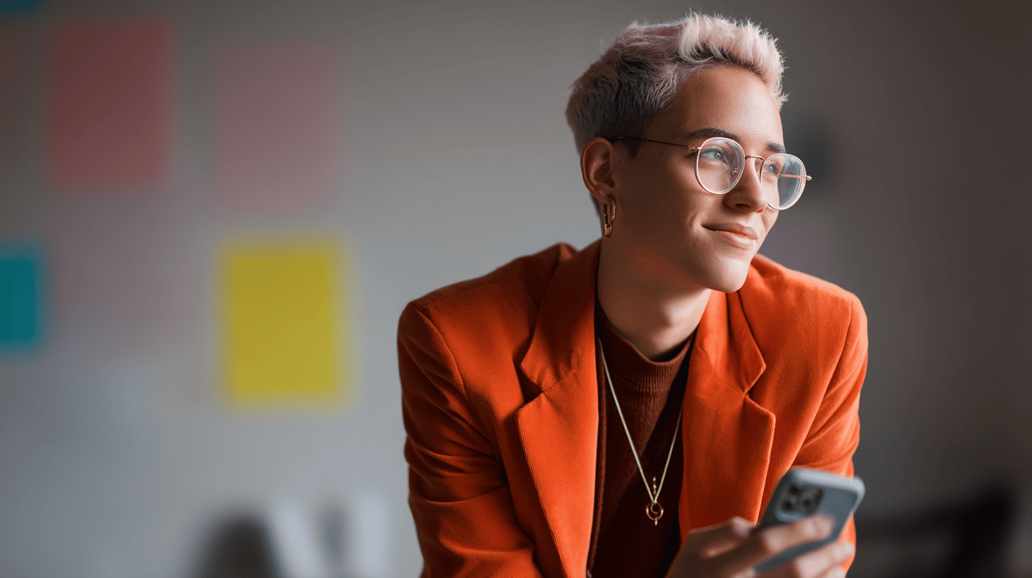 A person in an orange blazer, brown top, and glasses holds a phone, smiling, with colorful post-it notes in the background.