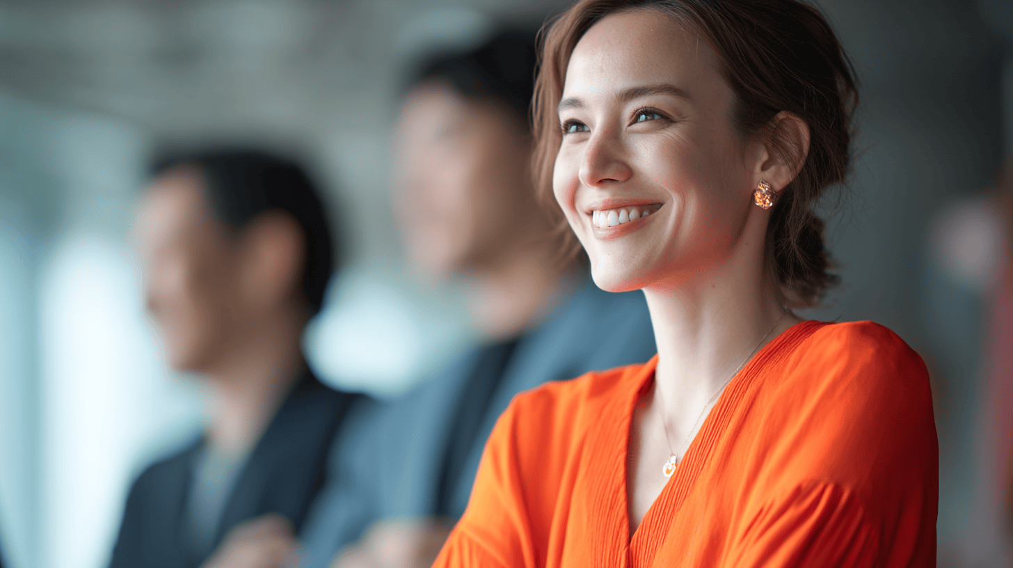A smiling woman in an orange shirt, with two blurred figures behind her, in a bright modern setting.
