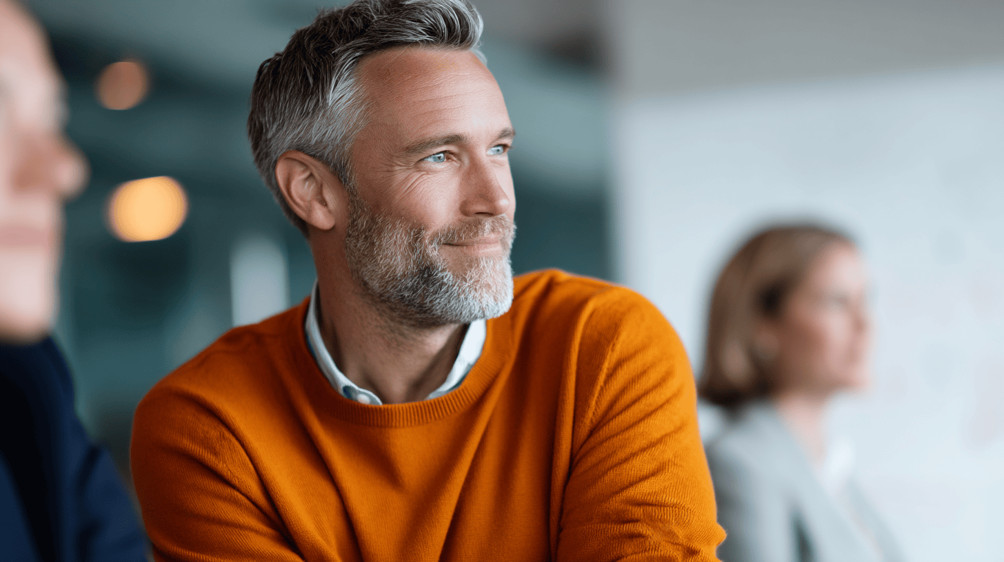 A man with gray hair and a beard wears an orange sweater, smiling while looking to the side, with two blurred people in the background.