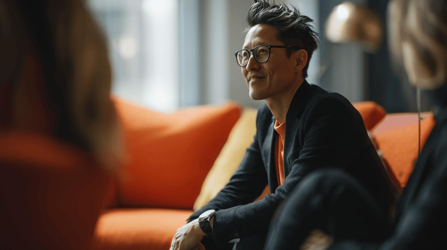 Man in glasses sitting on an orange couch, smiling while engaged in a conversation with another person.