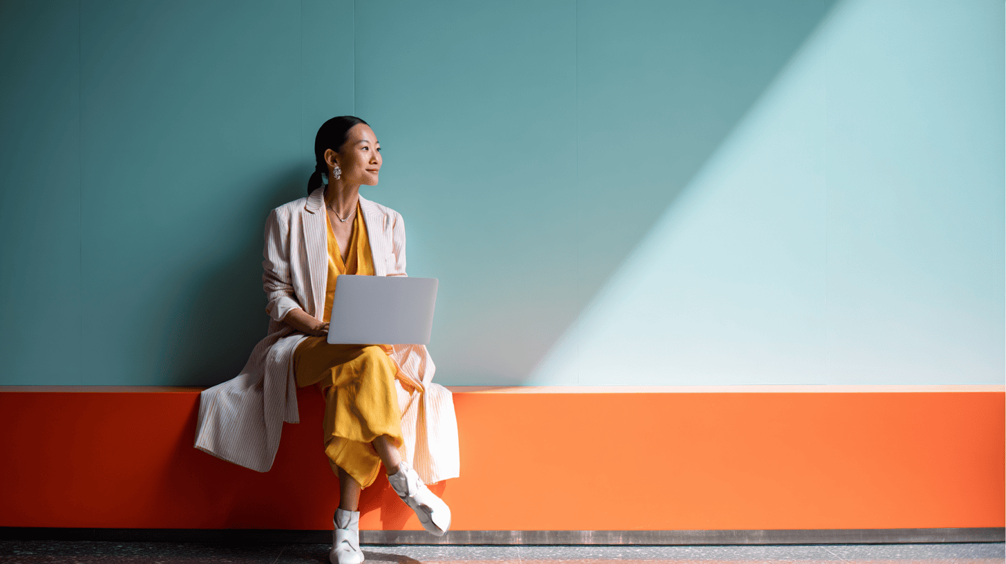 A woman sits on a colorful bench, working on a laptop in a bright, modern space with a teal wall and sunlight streaming in.