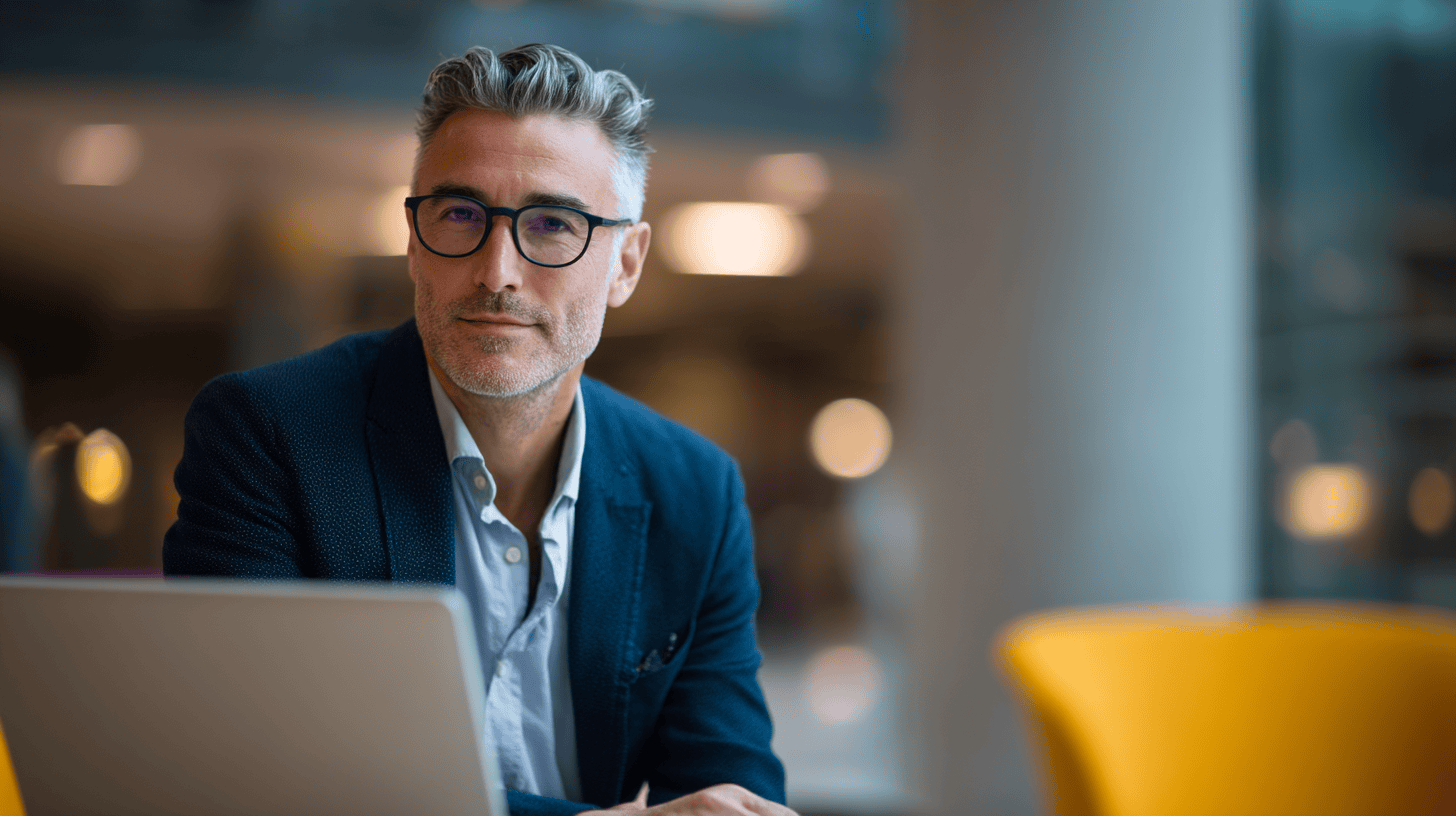 A man with gray hair and glasses sits at a laptop, wearing a blazer, in a modern, softly lit space.