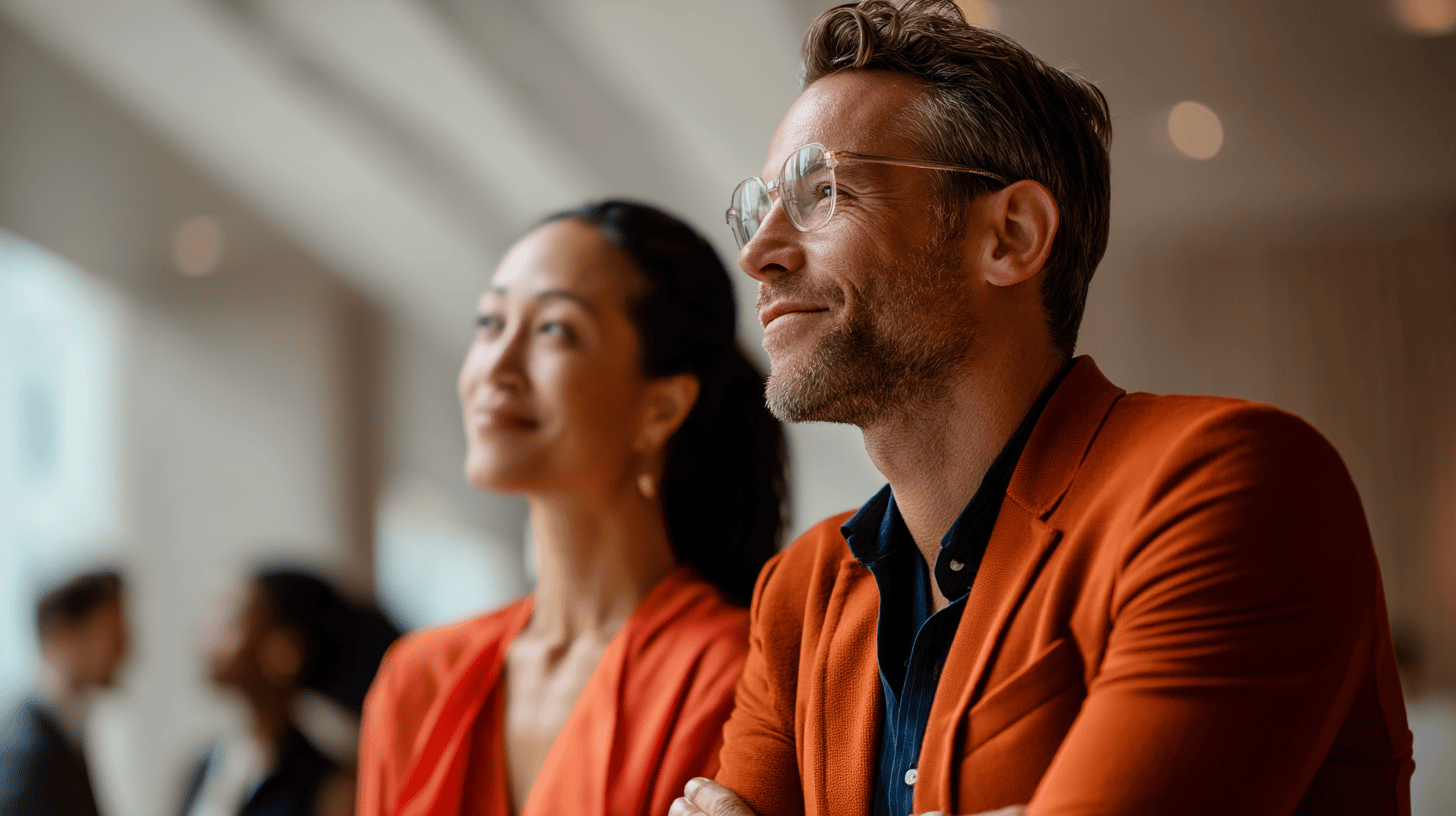 Two professionals, one in an orange blazer and glasses, and another in an orange top, sit facing forward, engaged.