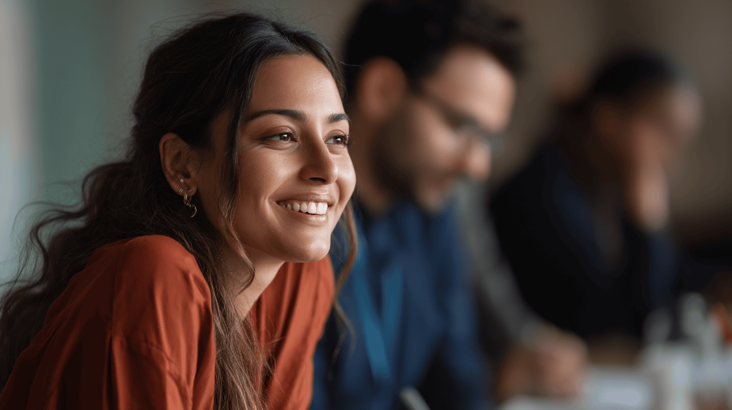 Smiling woman in an orange top at a meeting, with blurred colleagues in the background, all engaged in discussion.