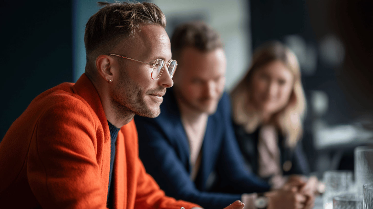 A man in an orange jacket sits at a table, with two blurred individuals behind him, in a modern meeting space.