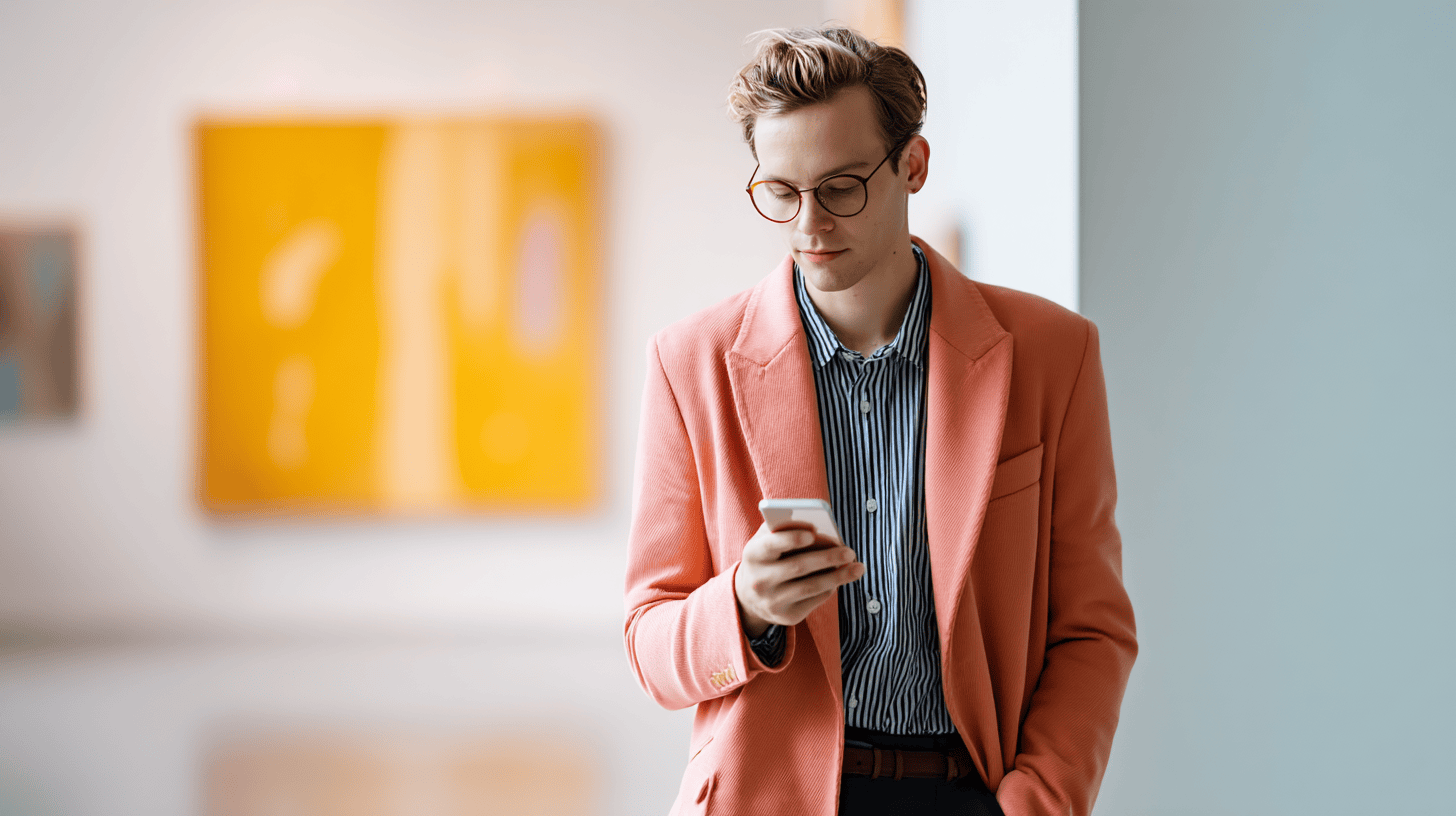 A young man in a coral blazer checks his phone in an art gallery with colorful abstract paintings in the background.