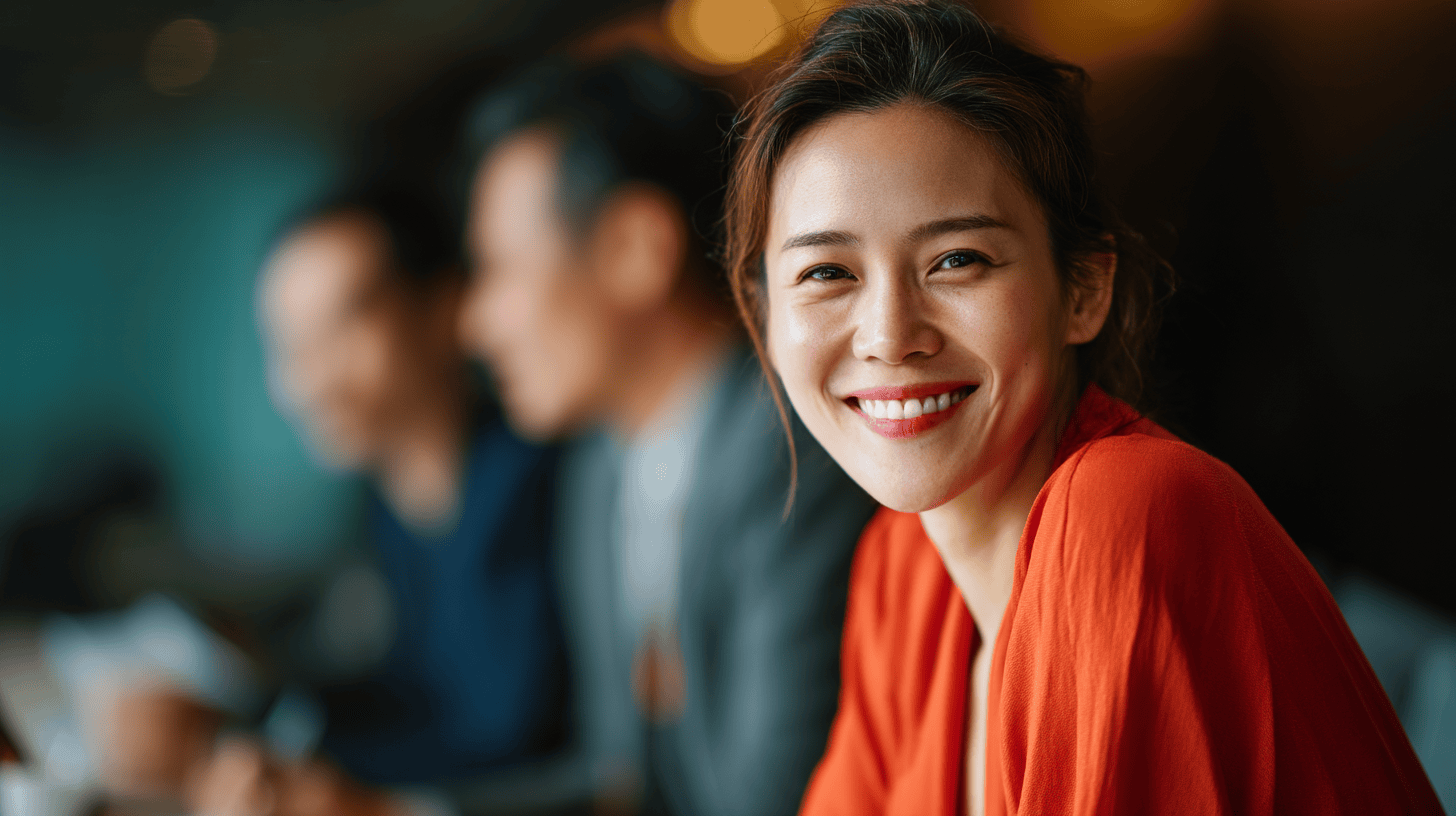 Smiling woman in a red top sits at a table with blurred figures of two men in the background, indoors.