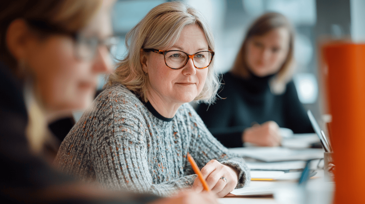 A woman in a sweater and glasses sits at a table with a pen, focused on a discussion, with two others blurred in the background.