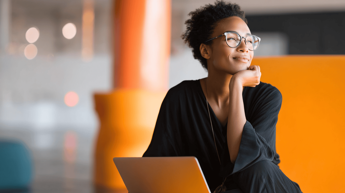 A woman in glasses smiles thoughtfully while sitting with a laptop in a modern, colorful workspace.
