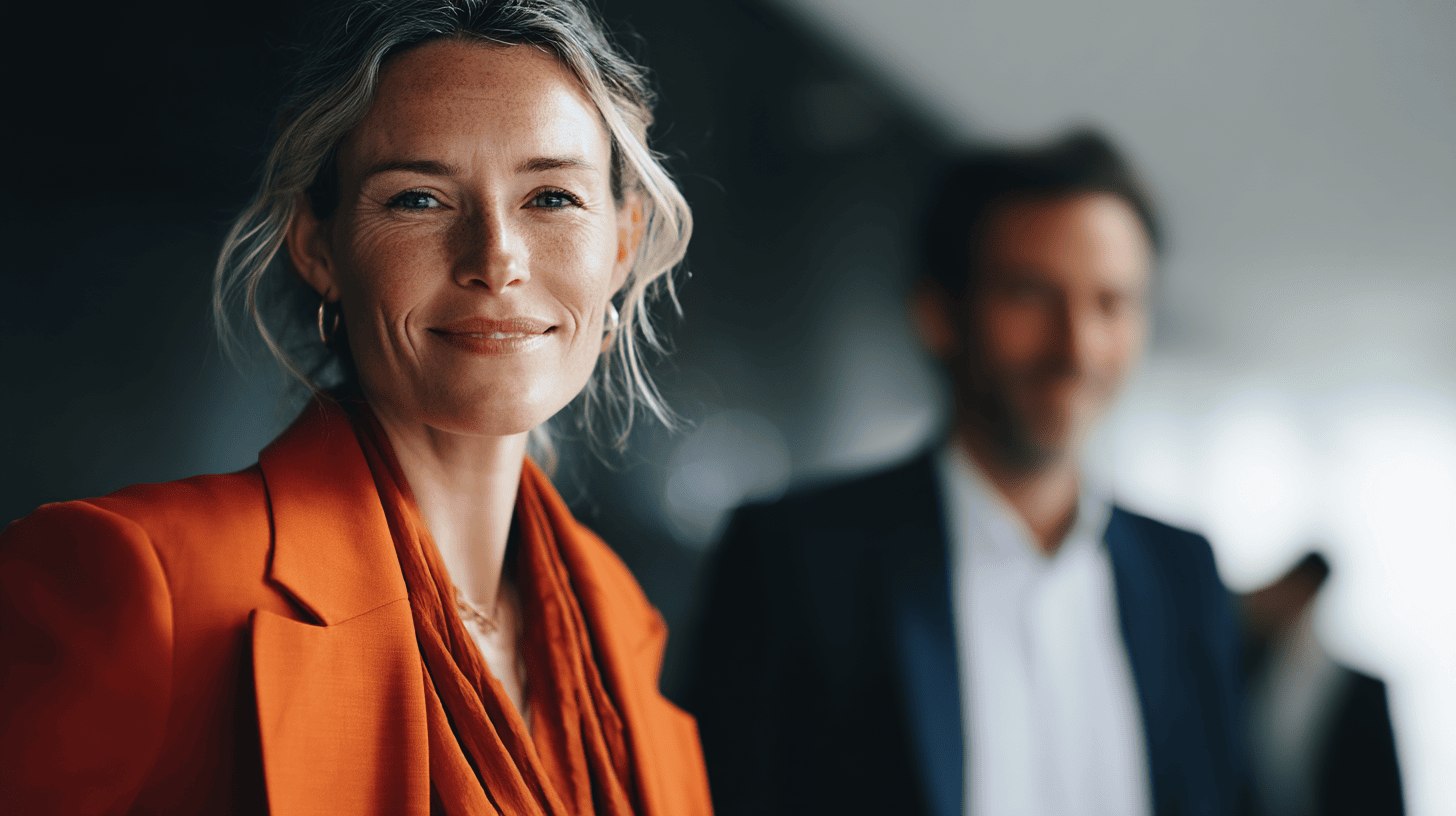 A woman in an orange blazer smiles, with a blurred man in a suit softly visible in the background.