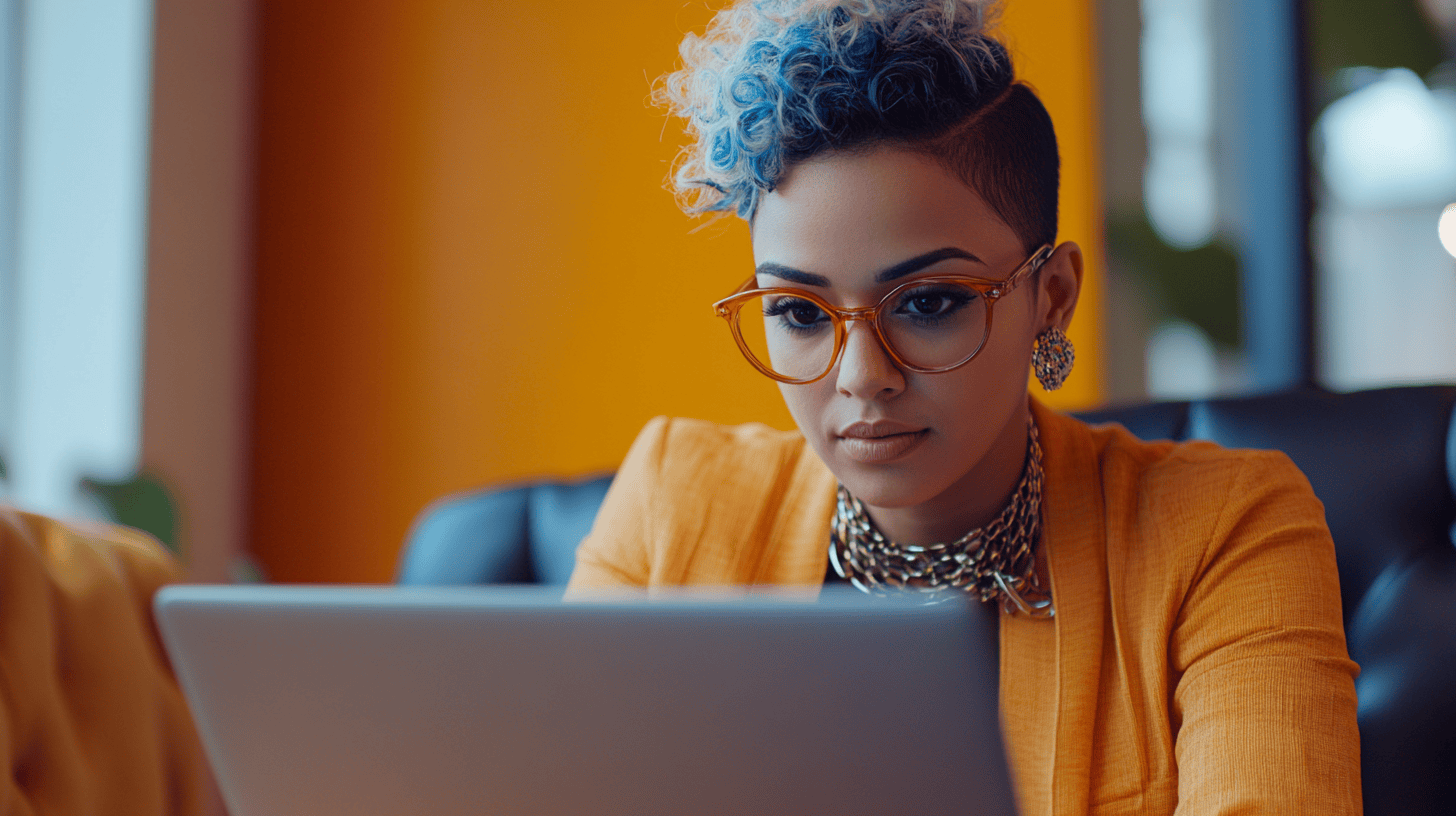 A woman with curly blue hair, orange glasses, and a yellow blazer focuses on a laptop in a bright indoor setting.