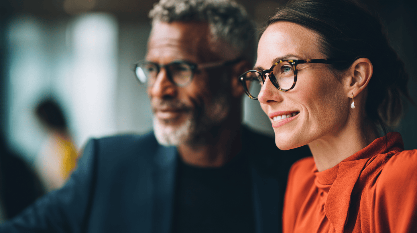 A close-up of a smiling woman in an orange blouse and glasses, alongside a man with glasses and a gray beard.