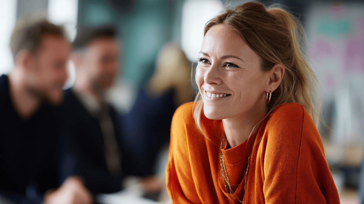 Smiling woman in an orange sweater, seated in a bright space with blurred coworkers in the background.
