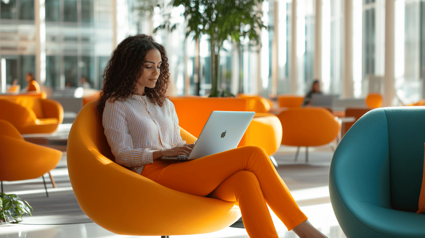 A woman in a striped shirt and orange pants sits in a modern chair, working on her laptop in a brightly lit, stylish space.