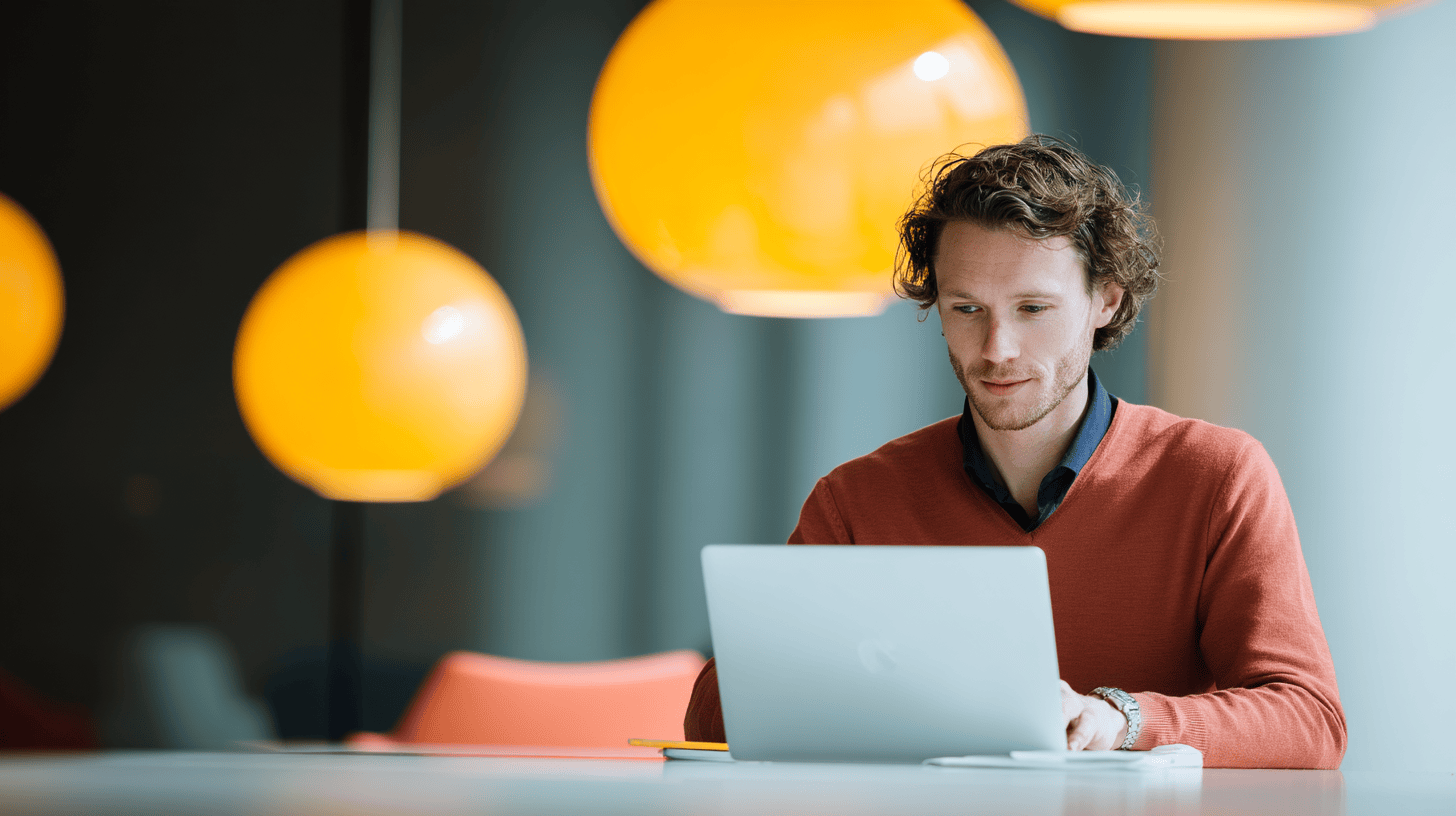 A man with curly hair works on a laptop in a modern space with orange pendant lights.