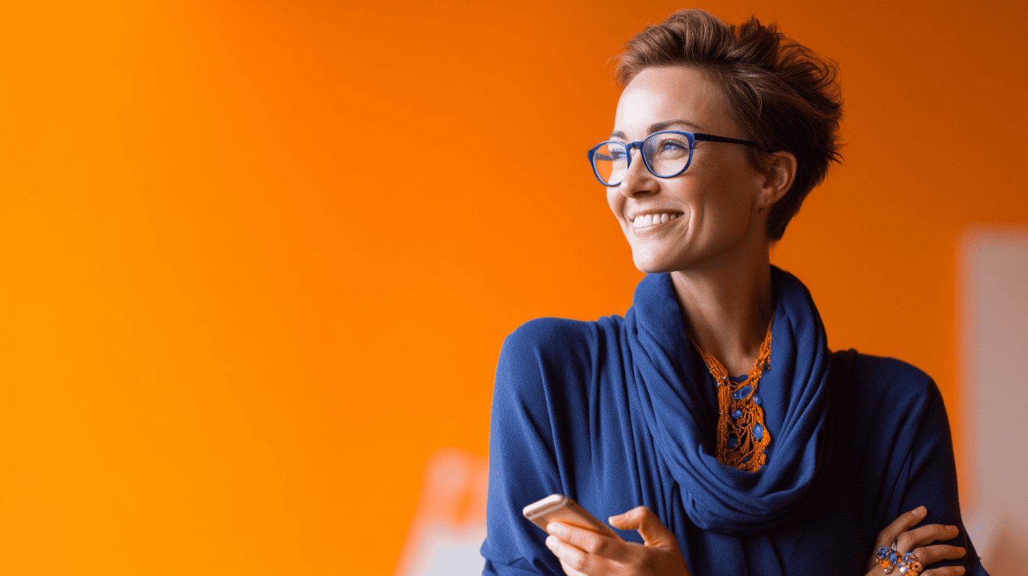 Smiling woman in glasses, wearing a blue sweater, holding a phone against a bright orange background.