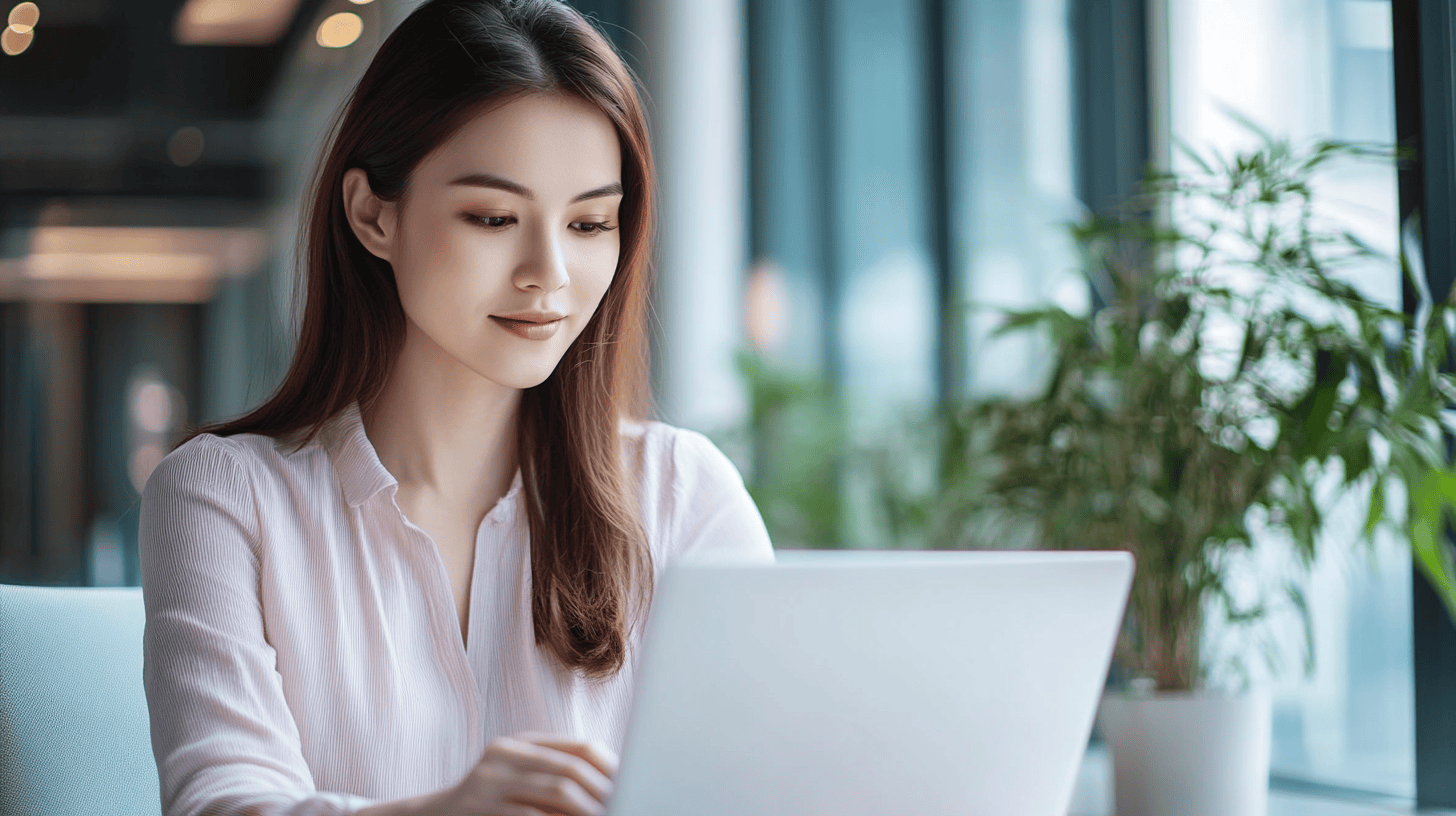 A woman in a light pink blouse works on a laptop near large windows with greenery in the background.