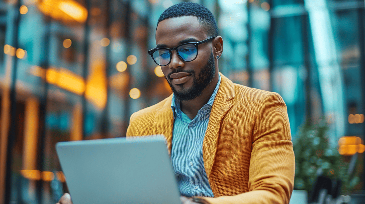 A man in a bright yellow blazer and blue glasses focuses on a laptop in a modern, well-lit space.