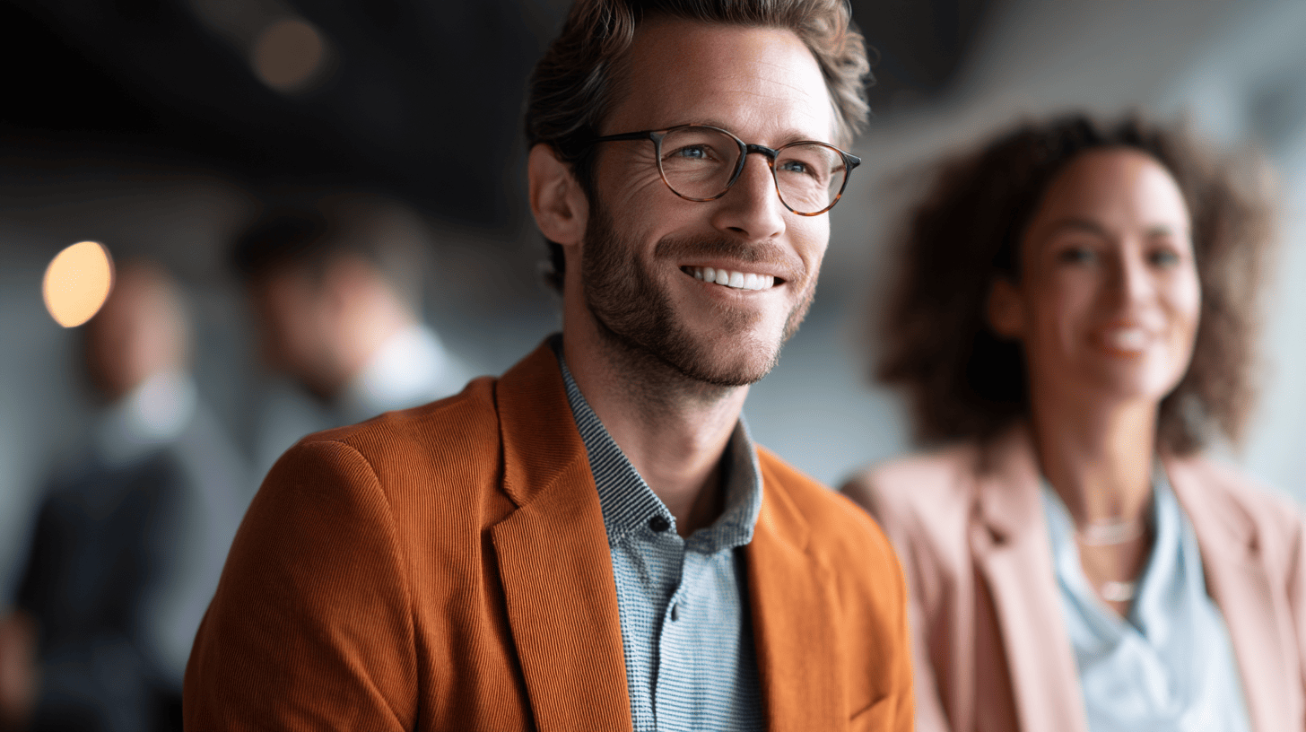 Smiling man in an orange blazer with a blurred woman in a pink suit behind him, set in a bright, modern space.