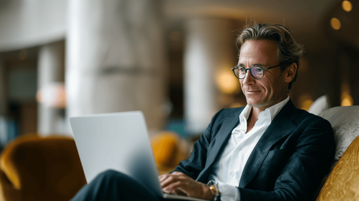 A man in a suit sits comfortably on a couch, working on a laptop in a stylish, modern interior.