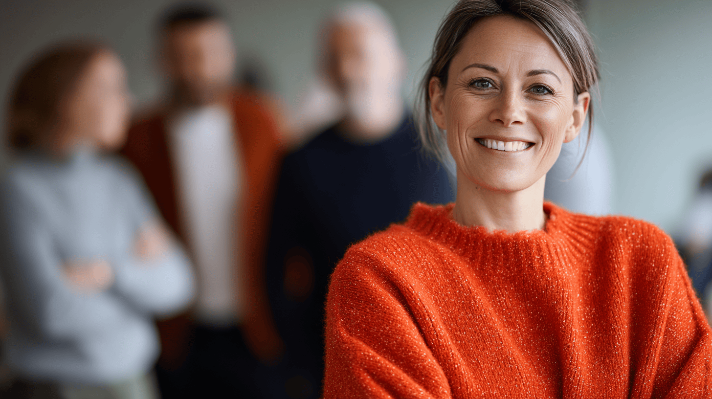 Smiling woman in an orange sweater, with blurred colleagues in the background, suggesting a collaborative atmosphere.