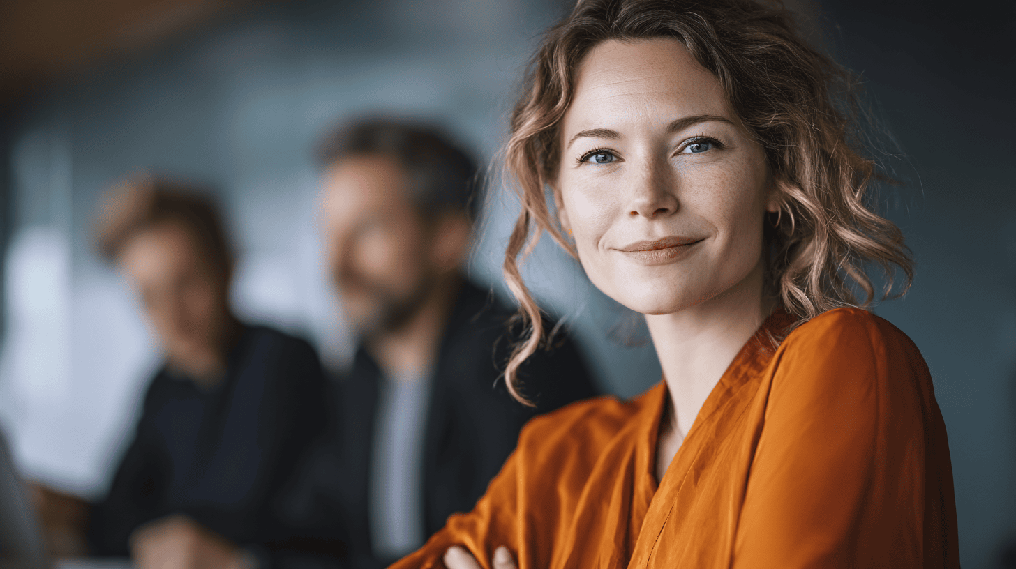 A smiling woman in an orange blouse sits at a table, with two blurred individuals in the background.