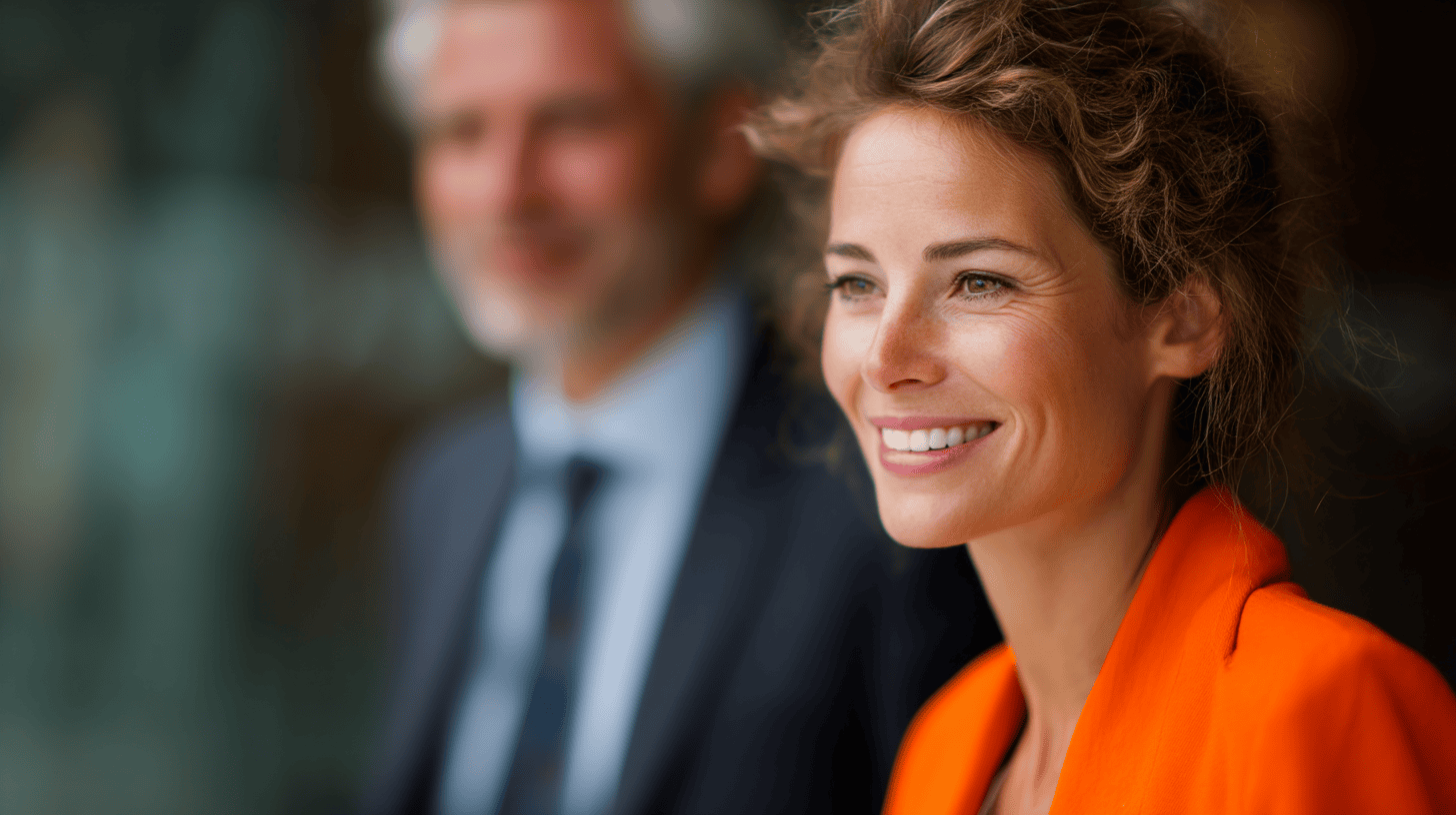 A woman in an orange blazer smiles warmly, with a blurred man in a suit slightly behind her, set against a soft background.