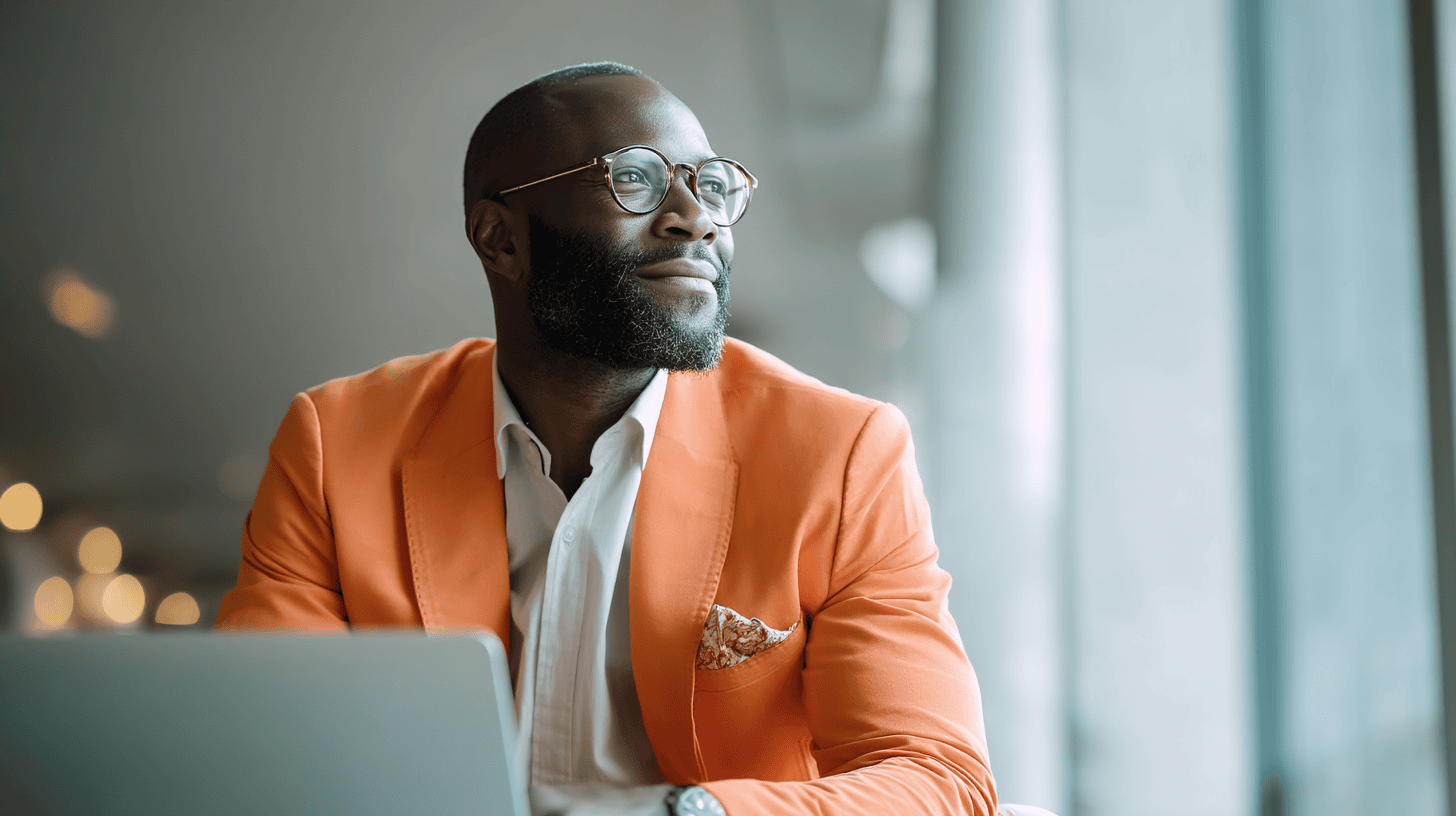 A man in an orange blazer and glasses gazes thoughtfully out a window, seated at a desk with a laptop.