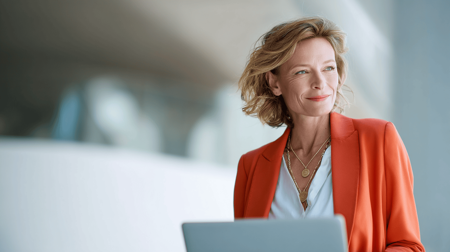 A smiling woman in an orange blazer holds a laptop, standing in a bright, modern space.