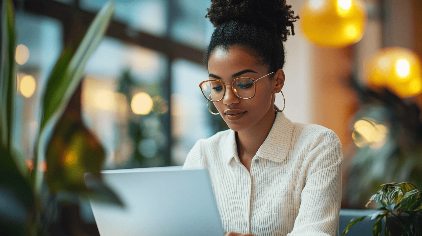 A woman with curly hair and glasses focuses on a laptop, surrounded by plants and soft, warm lighting.