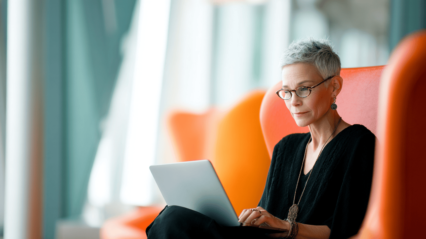 A woman with short hair and glasses sits in an orange chair, focused on her laptop in a modern workspace.