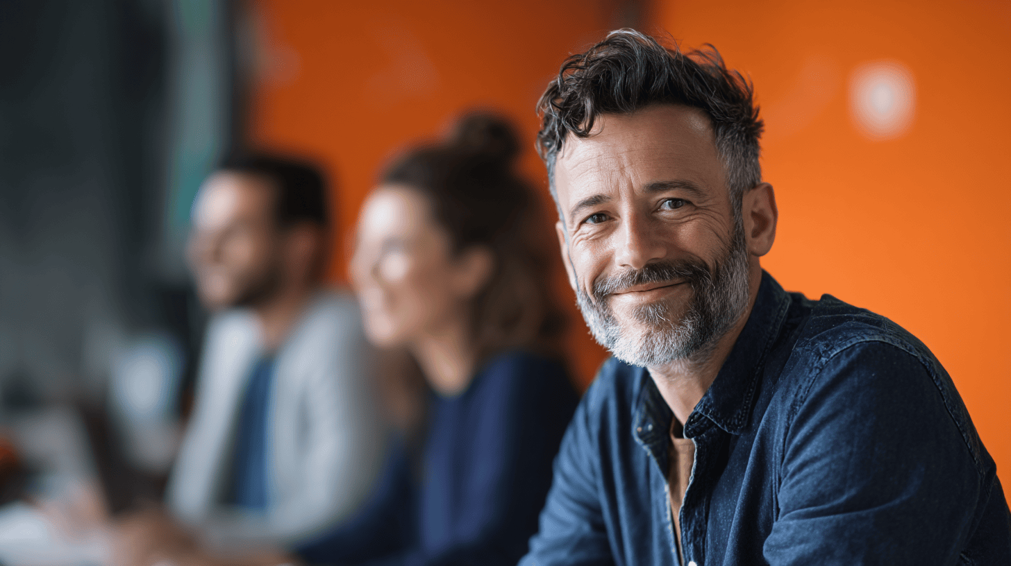 Smiling man with gray hair in a denim shirt sits at a table with blurred colleagues against an orange wall.