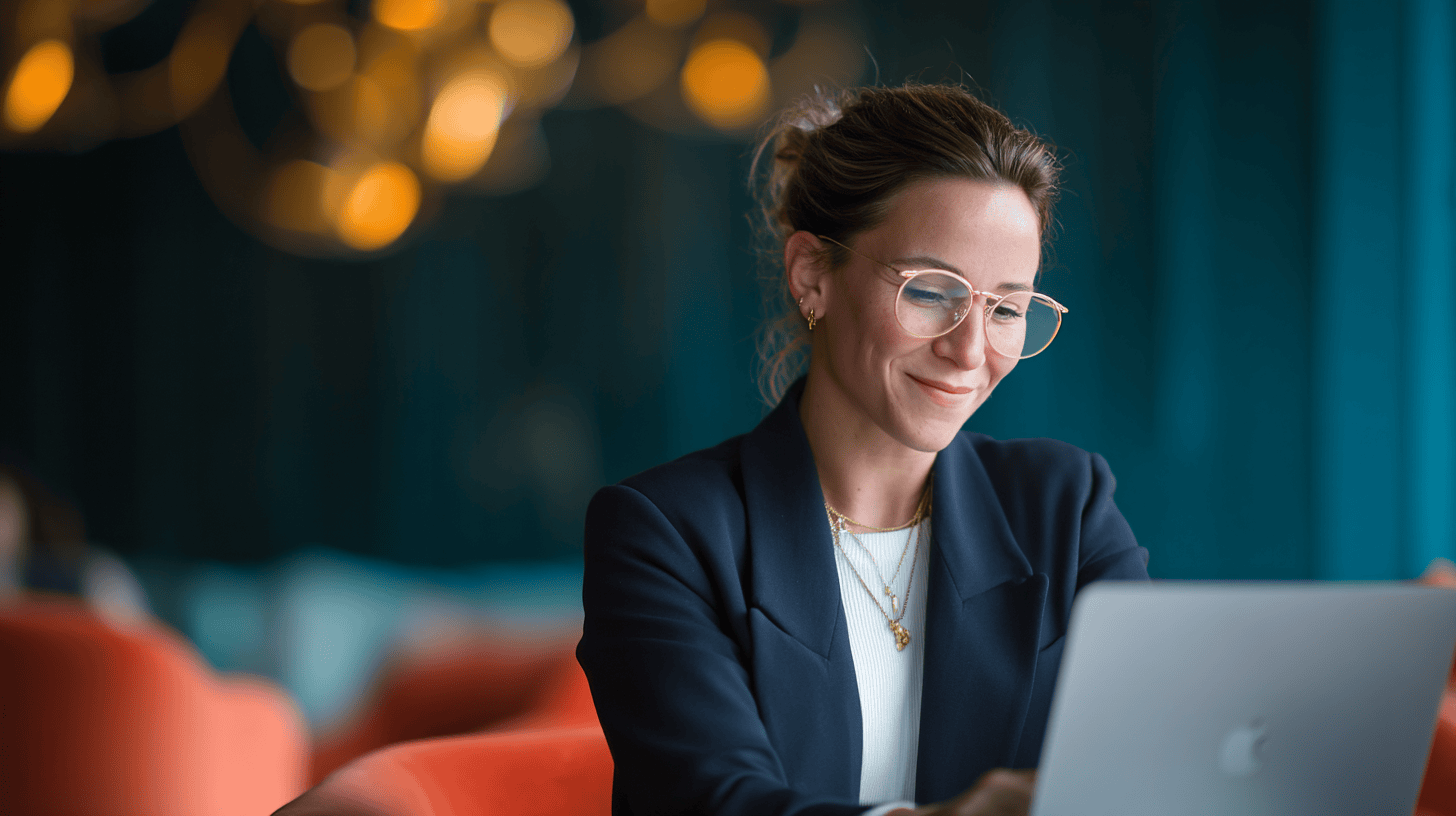 A woman in a black blazer smiles while working on a MacBook in a modern, cozy setting with orange and teal hues.