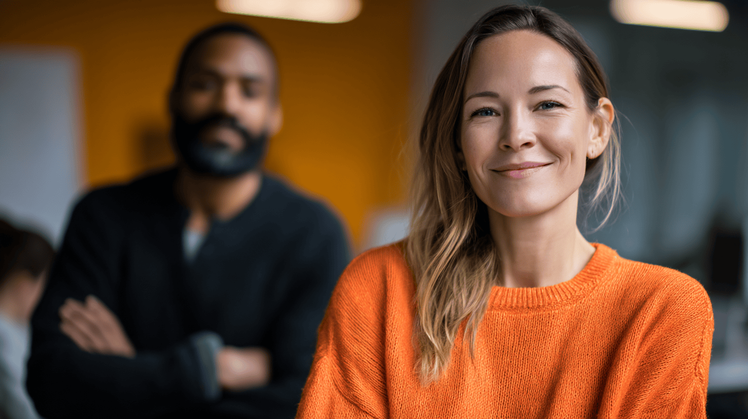A smiling woman in an orange sweater stands in the foreground, with a man in a black sweater blurred in the background.