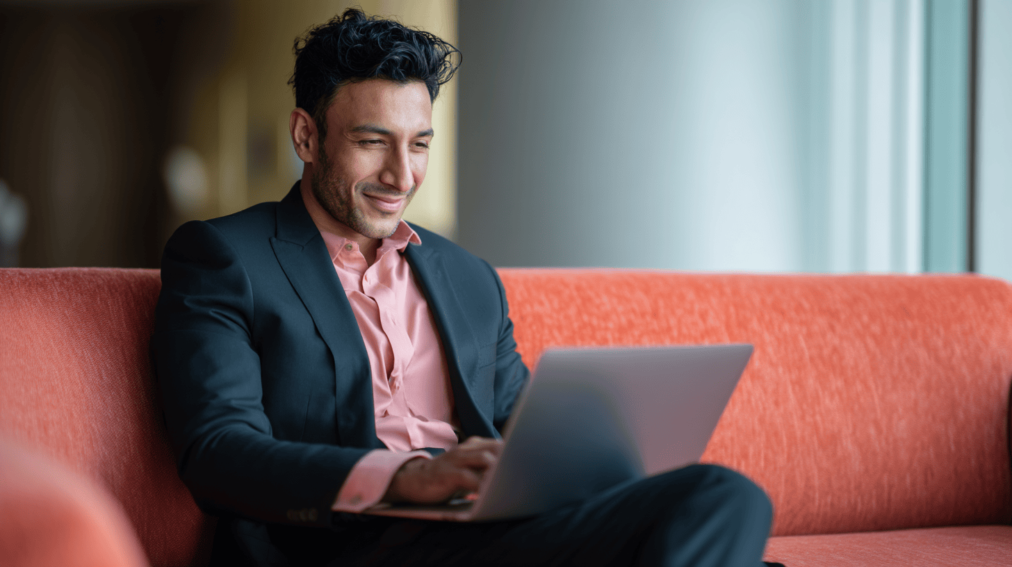 A man in a suit and pink shirt sits on an orange couch, working on a laptop with a smile.