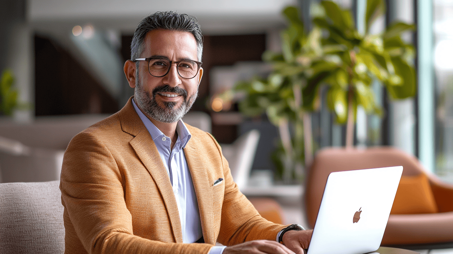 A smiling man in a tan blazer sits at a desk with a laptop, surrounded by greenery and modern furnishings.