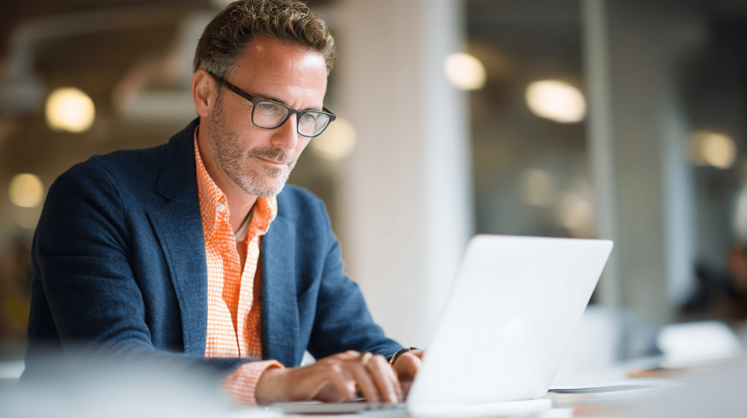 A focused man in a blazer and orange shirt working on a laptop in a modern office setting.