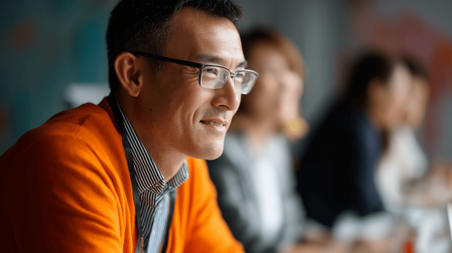 A smiling man in an orange sweater and glasses is seated at a table, with blurred colleagues in the background.