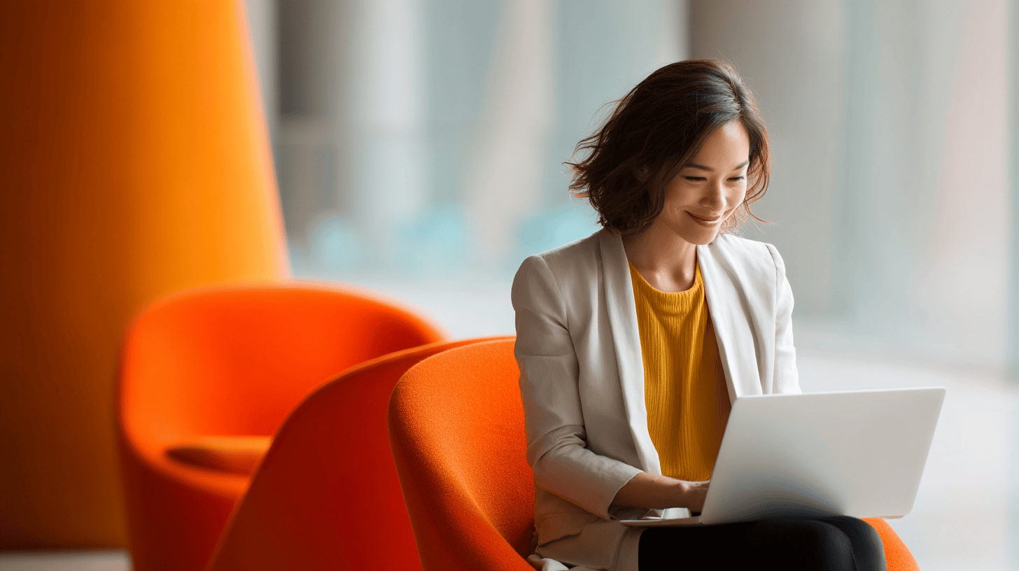 Smiling woman in a white blazer and yellow top sitting on an orange chair, using a laptop in a modern space.