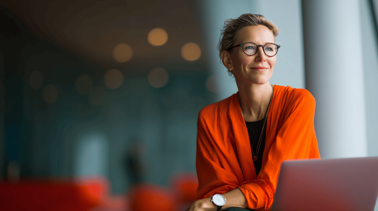 A woman with glasses and an orange jacket sits pensively beside a laptop in a softly lit modern setting.