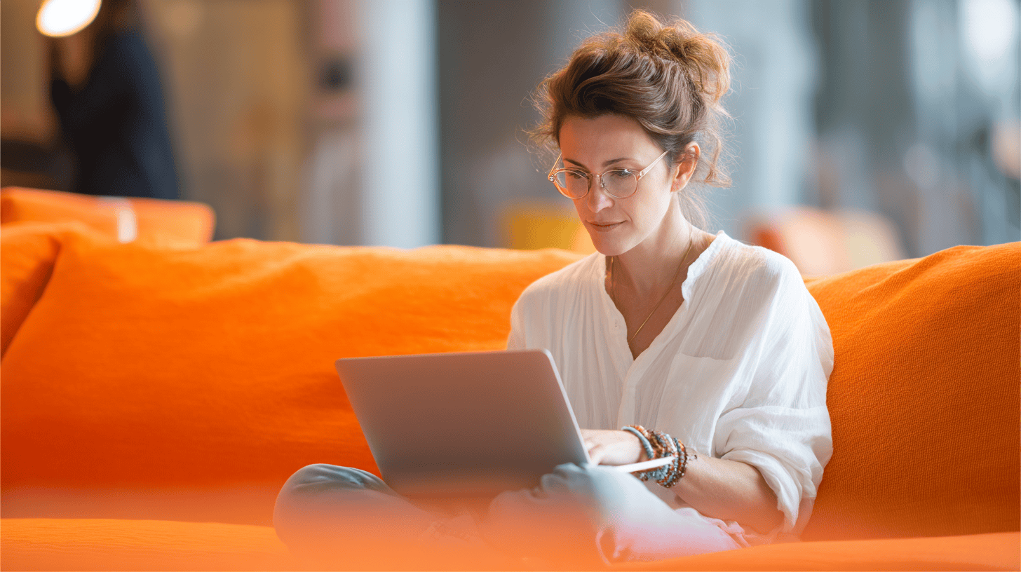 A woman sits on an orange couch, working on a laptop, wearing a white blouse and glasses, with bracelets on her wrist.