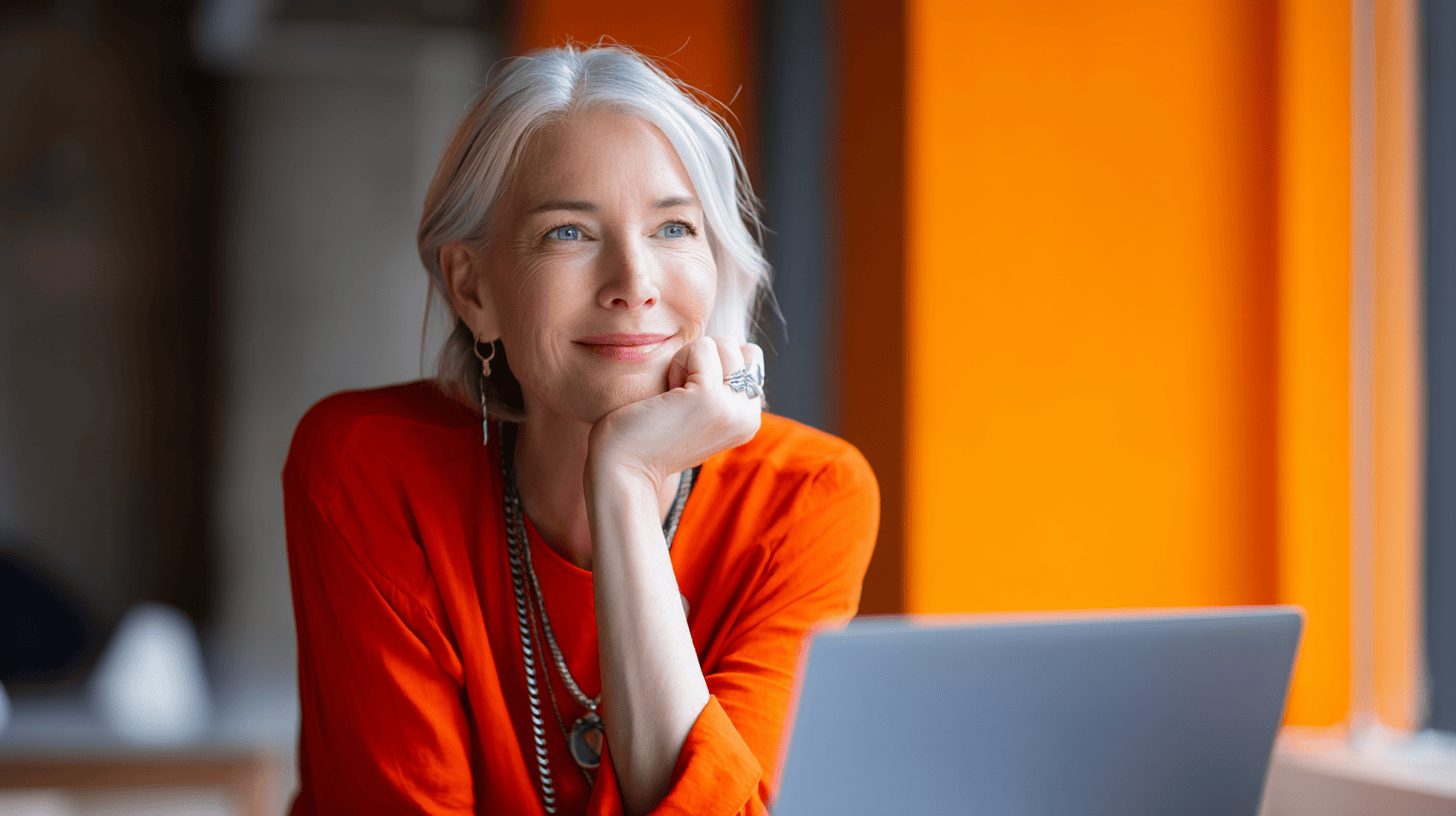 A smiling woman with gray hair rests her chin on her hand, wearing an orange shirt, with a laptop in front of her.