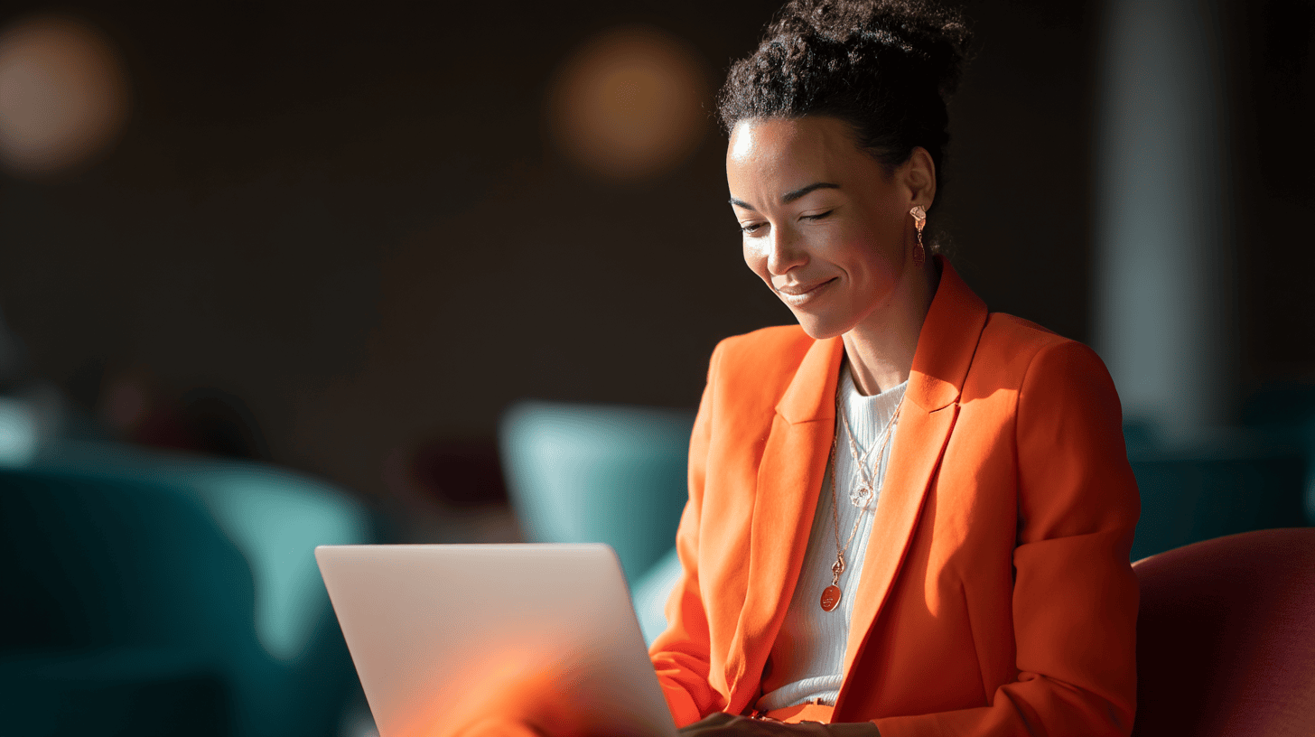 Smiling woman in an orange blazer using a laptop in a softly lit modern space with colorful seating.