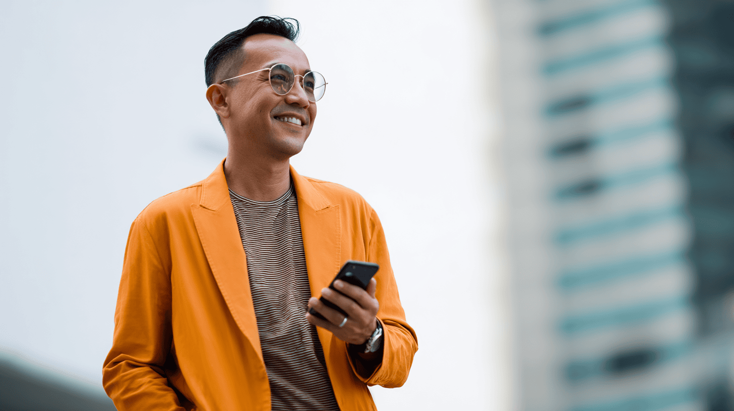 A man in an orange jacket and striped shirt smiles while holding a smartphone, with a blurred city skyline in the background.