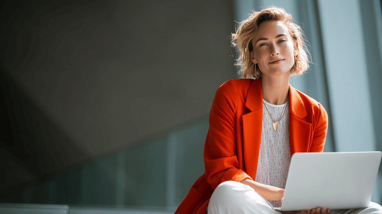 A woman in an orange blazer sits with a laptop, smiling confidently in a modern, bright space.