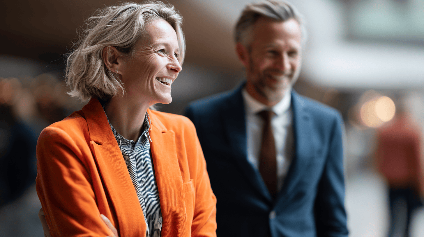 A smiling woman in an orange blazer and a man in a dark suit stand together, engaging in conversation indoors.