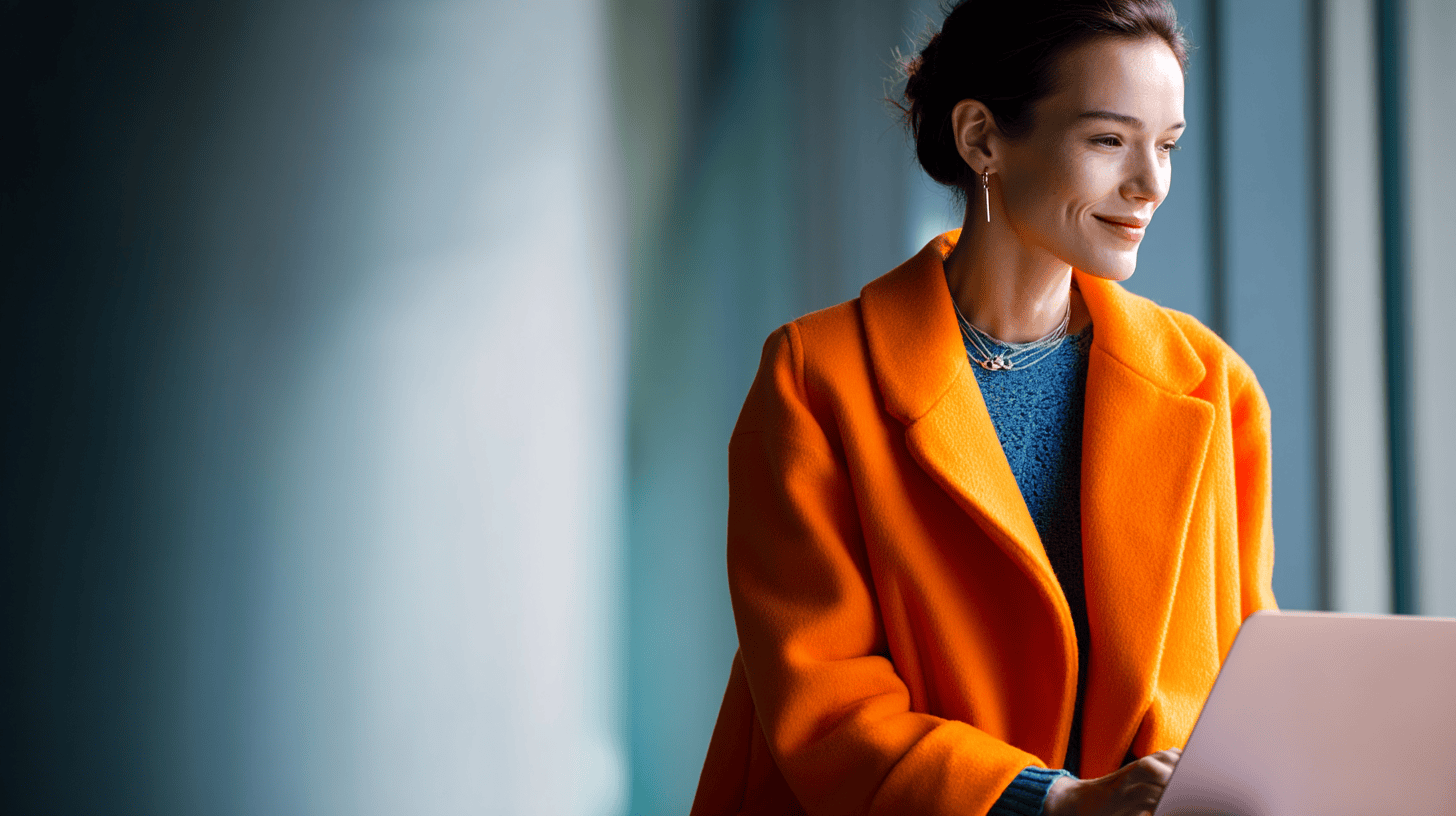 Woman in an orange coat smiles while using a laptop, with large windows in the background casting soft light.