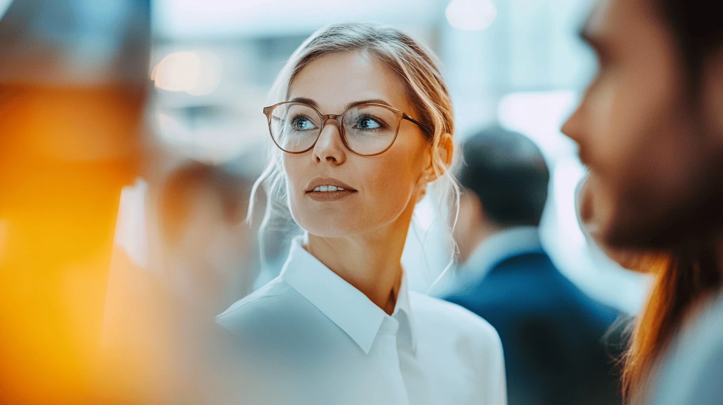 A professional woman in glasses looks thoughtfully while engaged in a conversation at a business event.